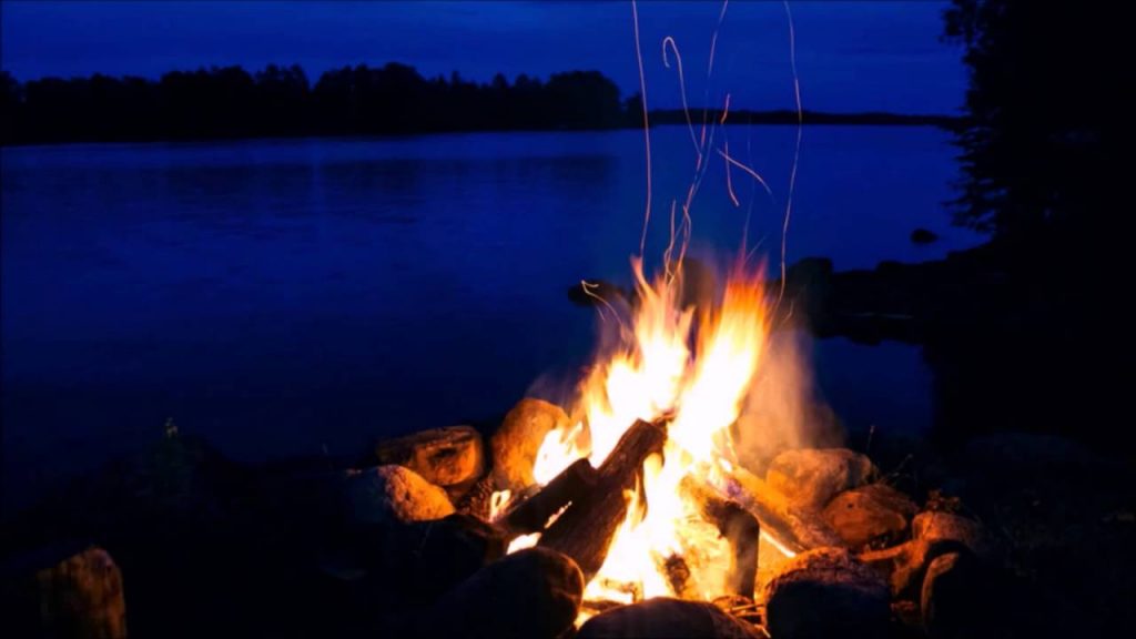 A long exposure of an open fire in a campground at night, with a lake in the background.