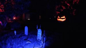 Halloween 2019 home at night, blacklight on tombstones, skeletal hands in the gorund, and a sleepy hollow pumpkin watching from the mailbox.
