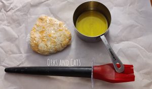 A small piece of wrapped dough, a measuring cup with olive oil, and a silicone basting brush resting on parchment paper.